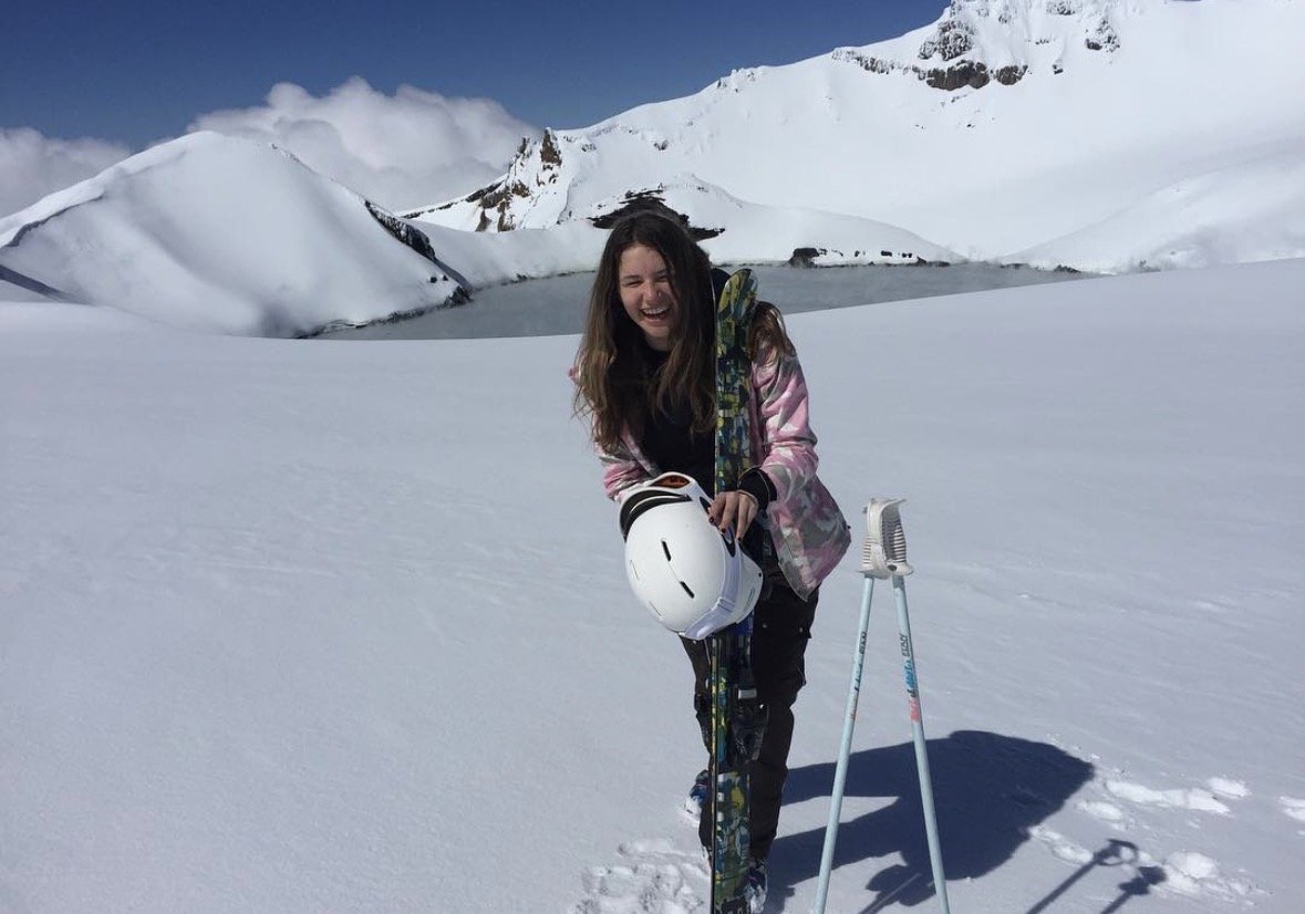 A young woman on a snowy mountain, smiling, holding a ski helmet, with skis and ski poles. Snow-covered mountains and a clear sky are in the background.