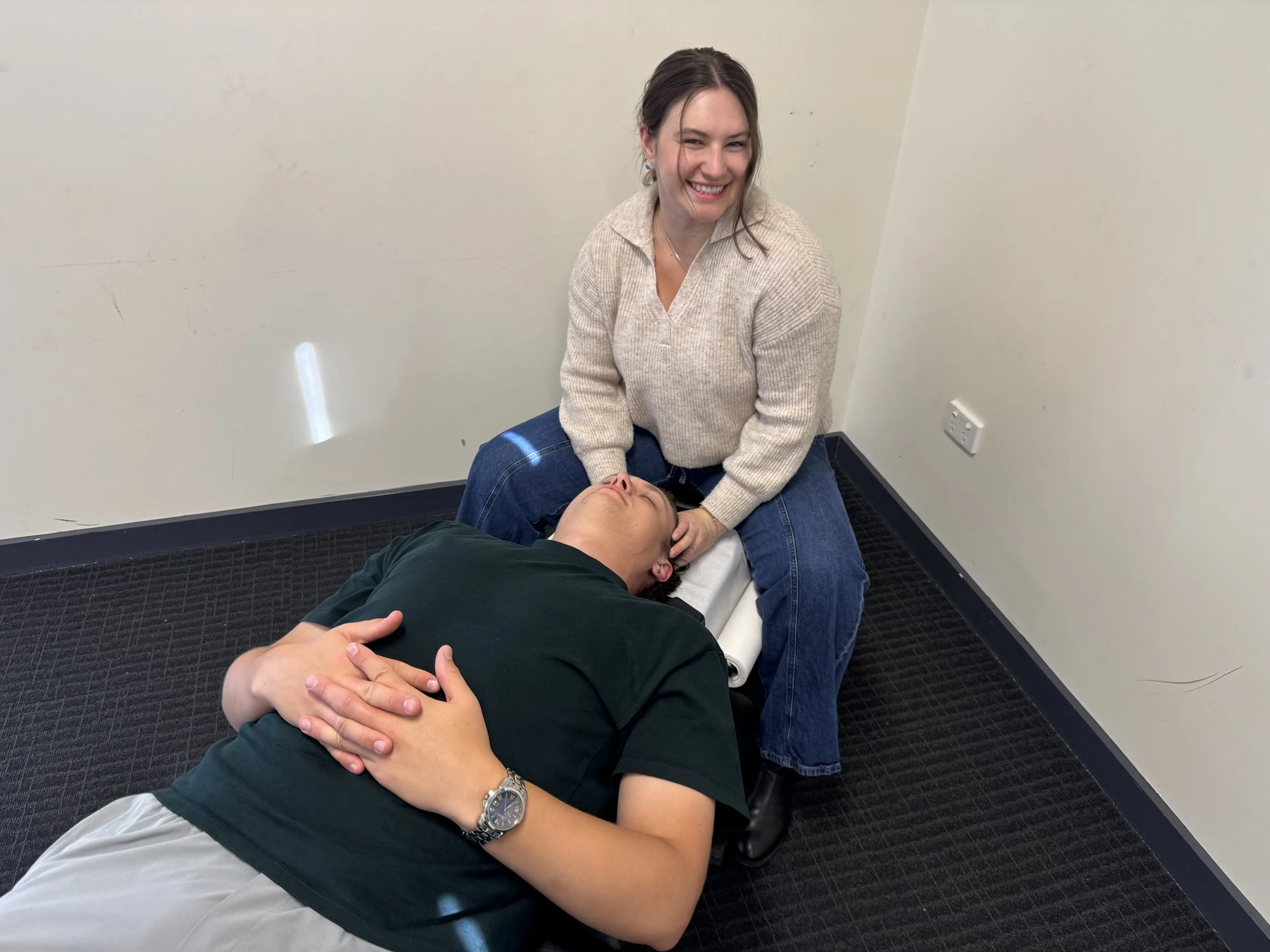 A woman smiling and sitting on the floor with a person lying down, holding their head, in a plain room.