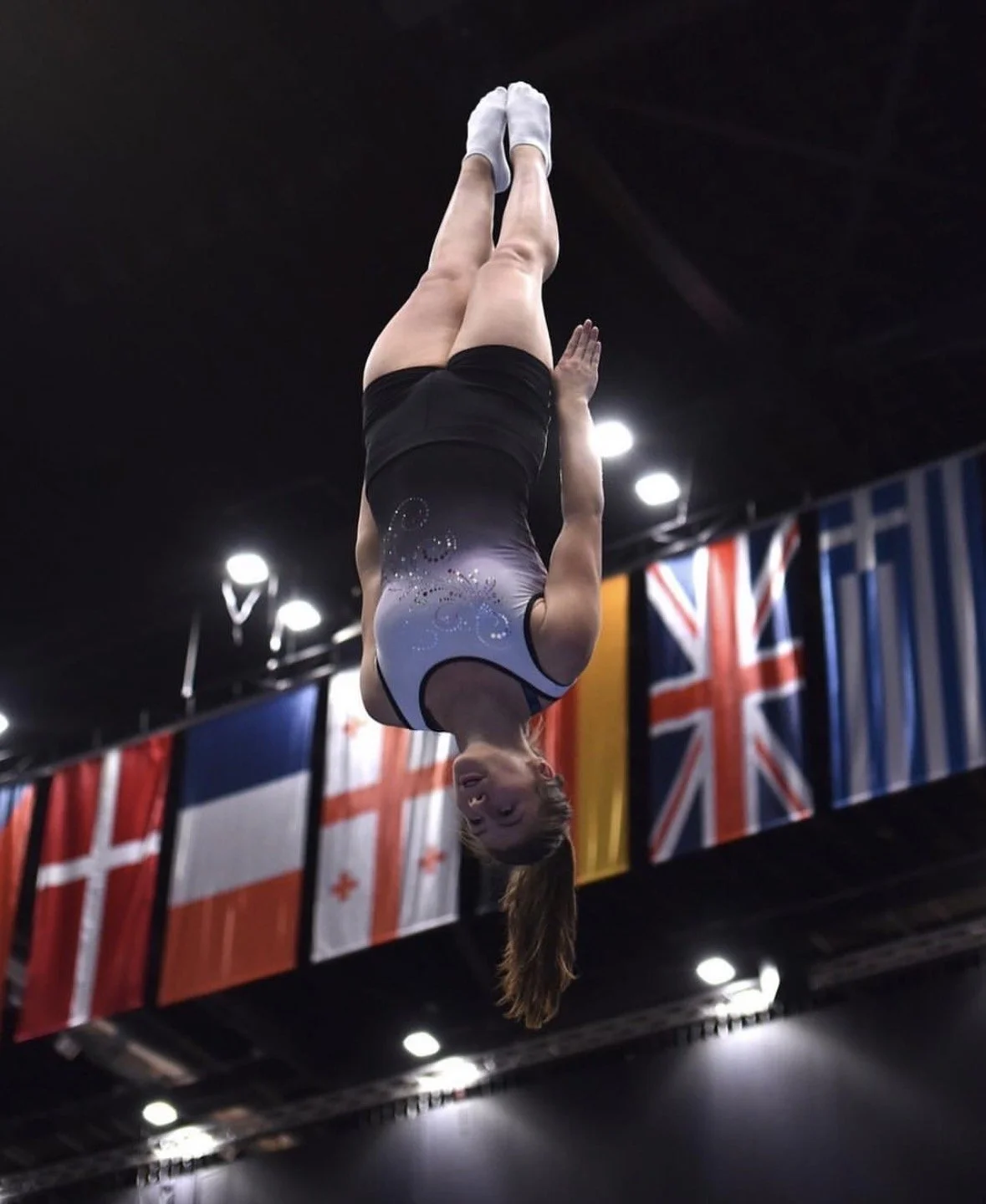 A gymnast in a black and grey leotard performing a flip or jump in an arena with flags from various countries hanging in the background.