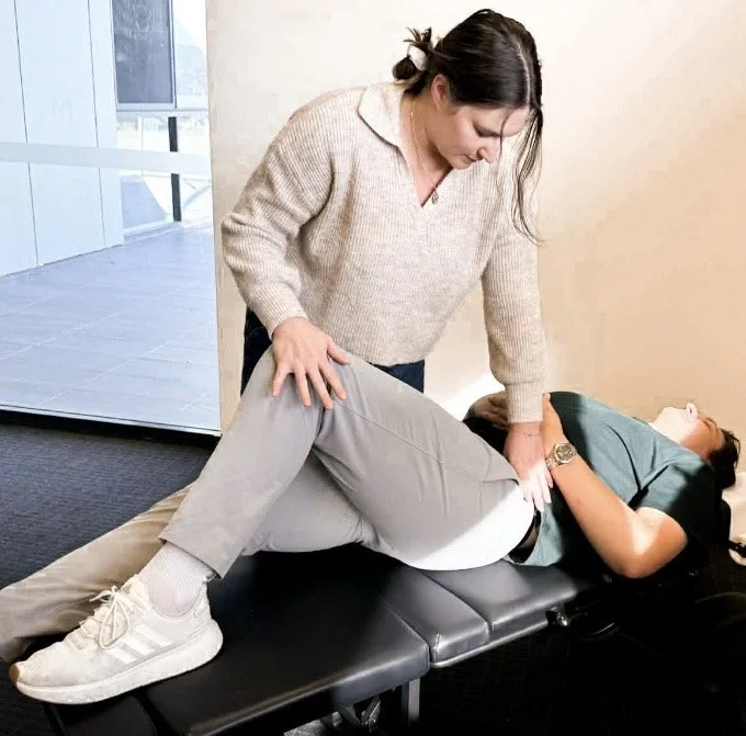 A woman in a beige sweater performing a chiropractic adjustment on a woman lying on a treatment table.