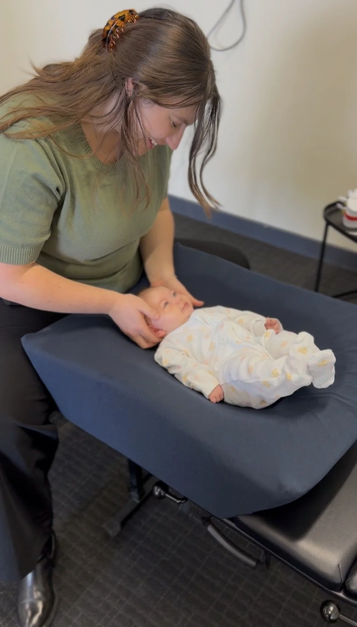A woman with a tortoise hair clip smiling while gently holding the head of a sleeping baby lying on a padded table.