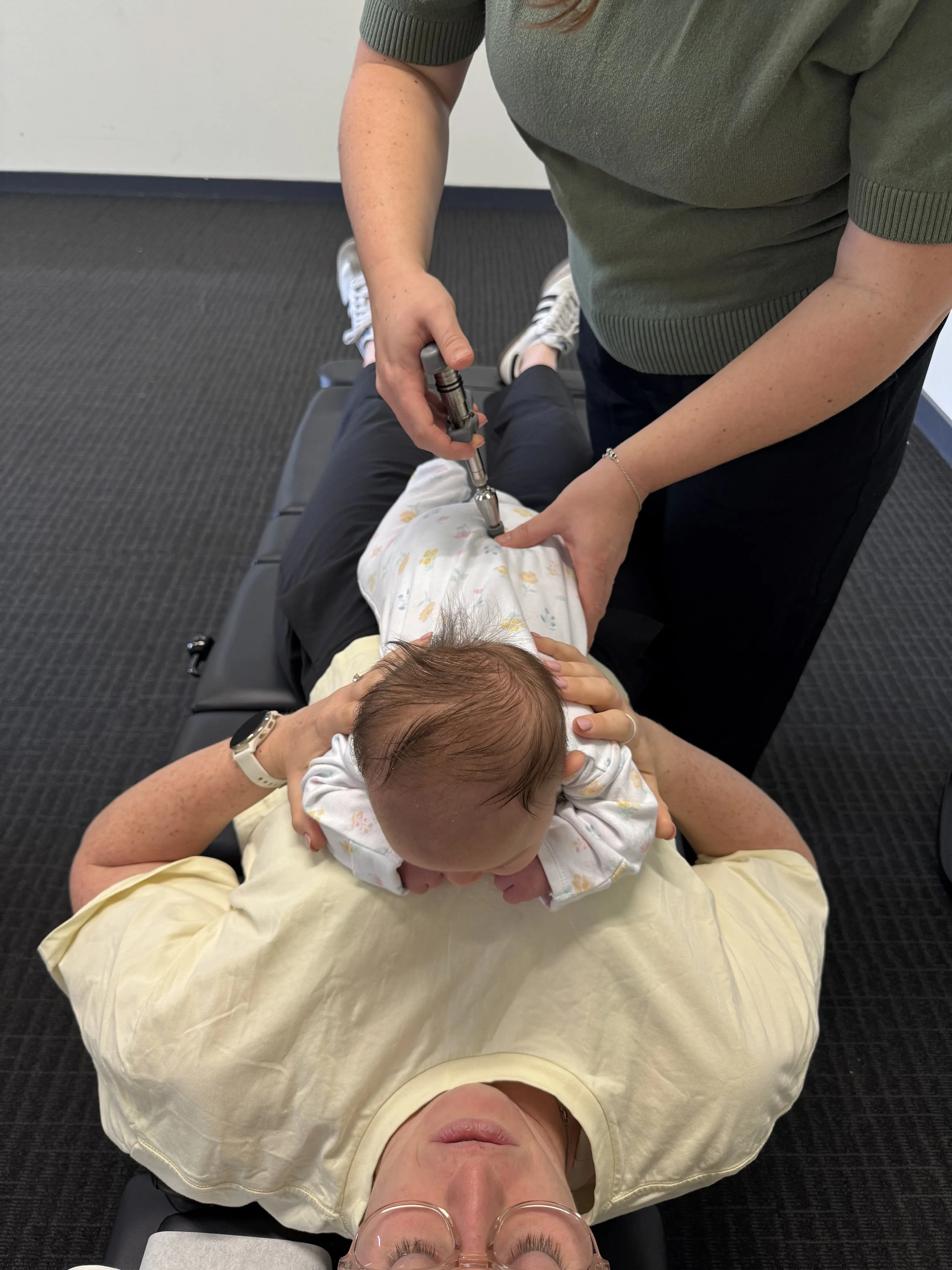 A person lying on their back on a medical treatment table with a young child on their stomach. A healthcare worker is administering a medical injection into the child's arm.