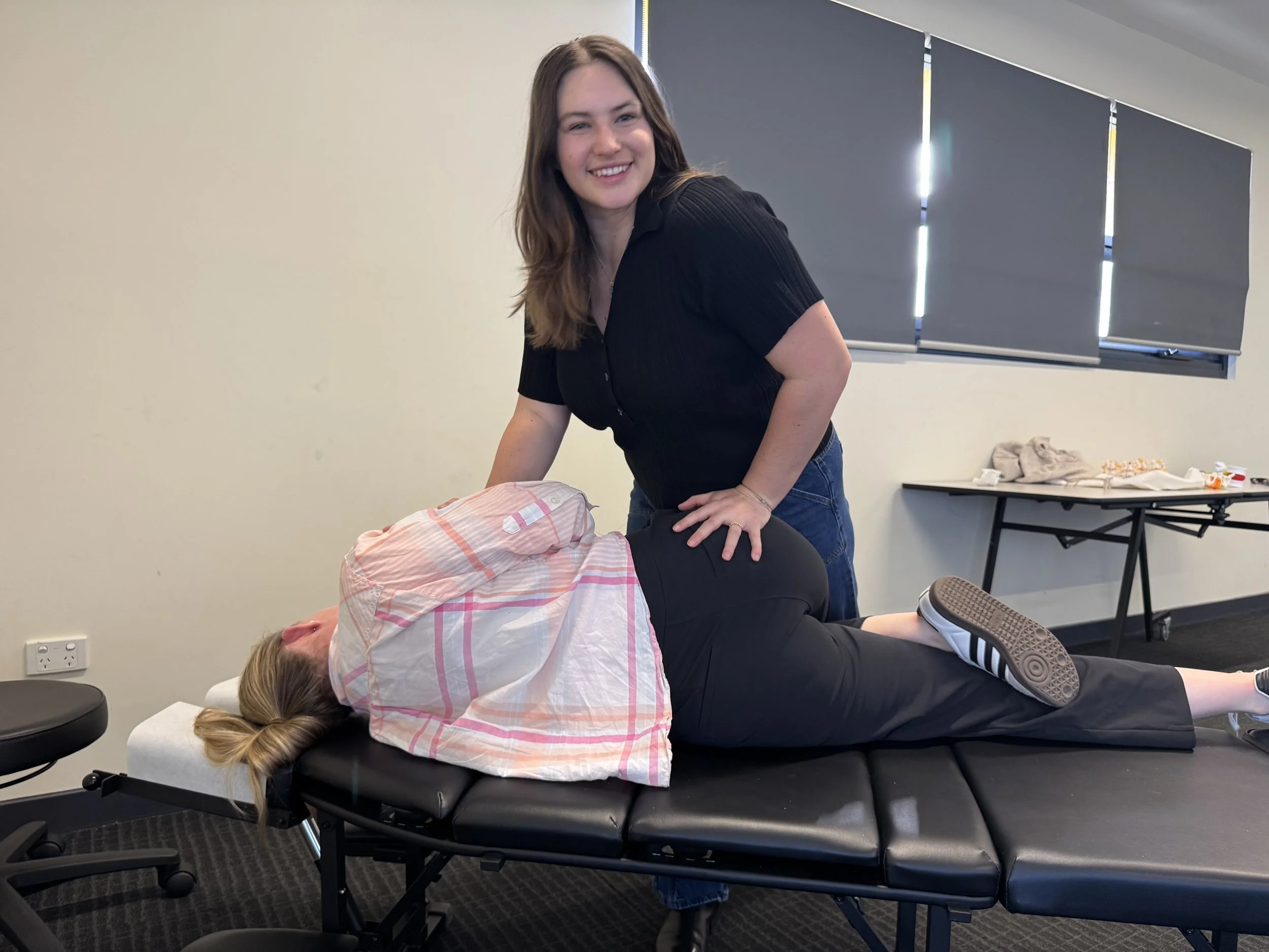 A woman getting a massage on her back while lying face down on a massage table, with another woman standing beside her in a therapy room.