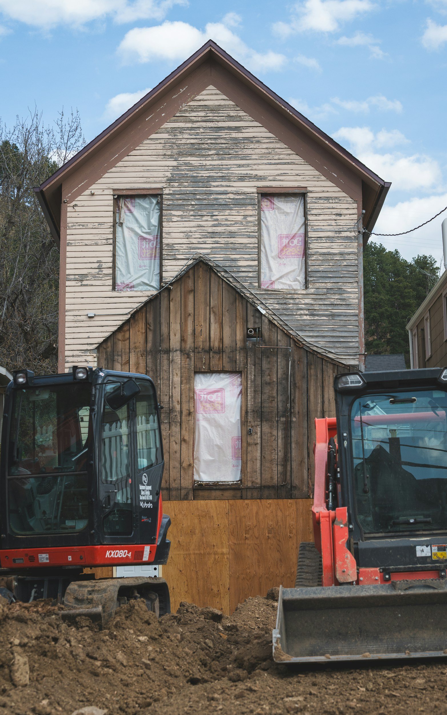 Construction site with two small excavators in front of a house that is being renovated, with the upper part of the house showing worn, peeling paint and covered windows, and the lower part covered in new wood.