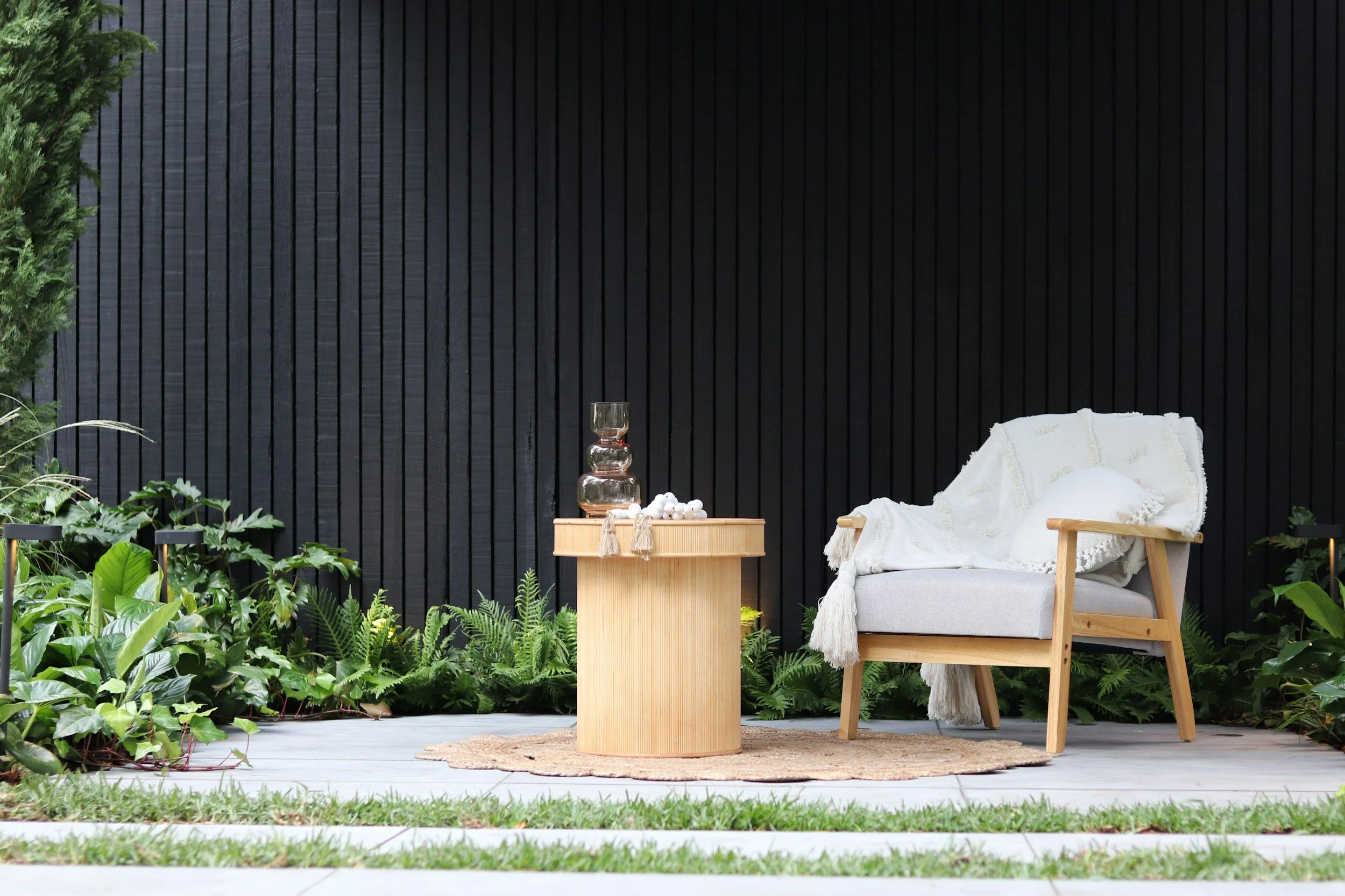 Outdoor patio with a white cushioned chair draped with a white blanket, a round natural fiber rug, a wooden side table with decorative objects, and lush green plants against a black vertical wooden fence.