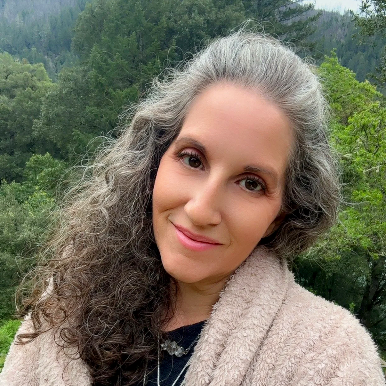 A woman with long, curly gray hair smiling outdoors with a background of green trees and mountains.
