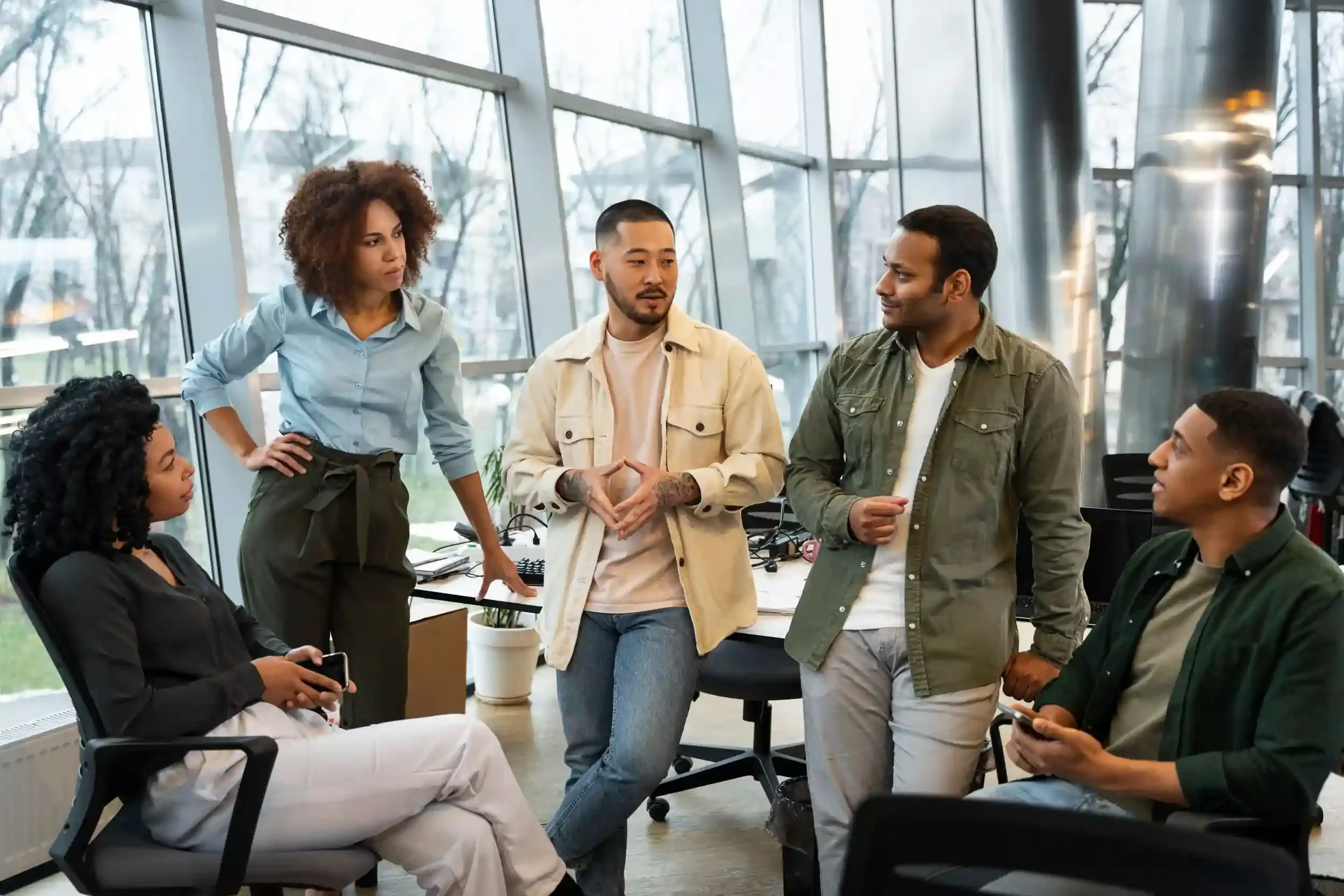 A diverse group of five young adults having a discussion in a modern office with large windows.