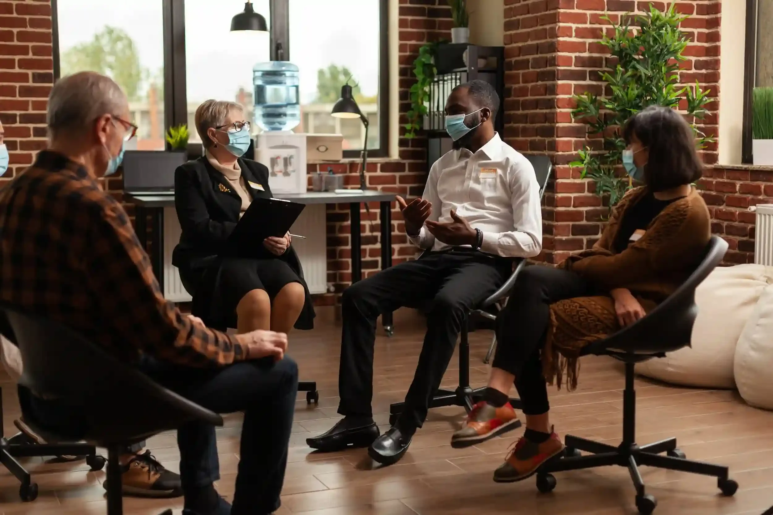 A diverse group of five adults, all wearing face masks, seated in a semi-circle in a modern office with brick walls, engaged in a discussion.