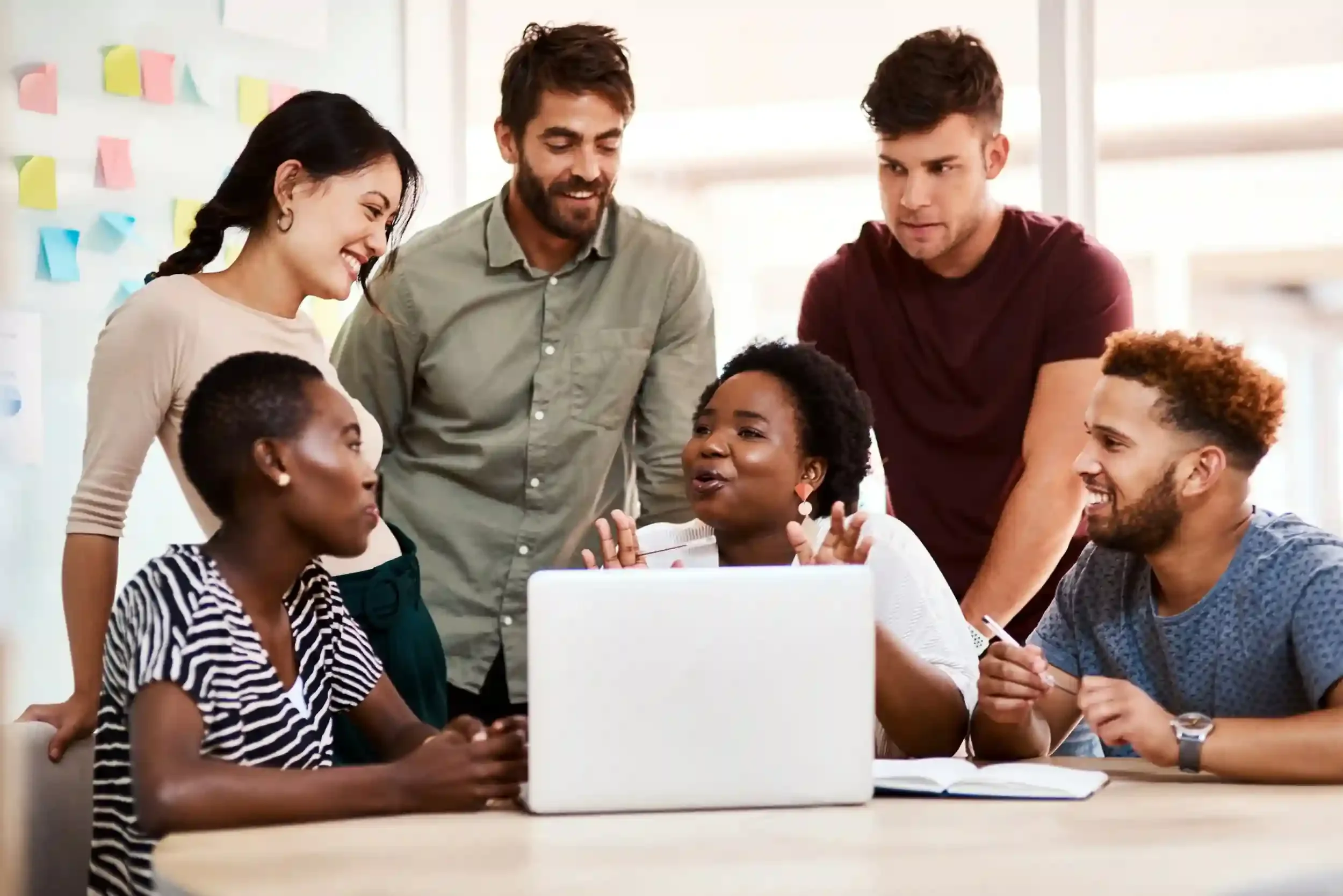 A diverse group of six people gathered around a laptop, engaged in a discussion or collaboration in an office setting.