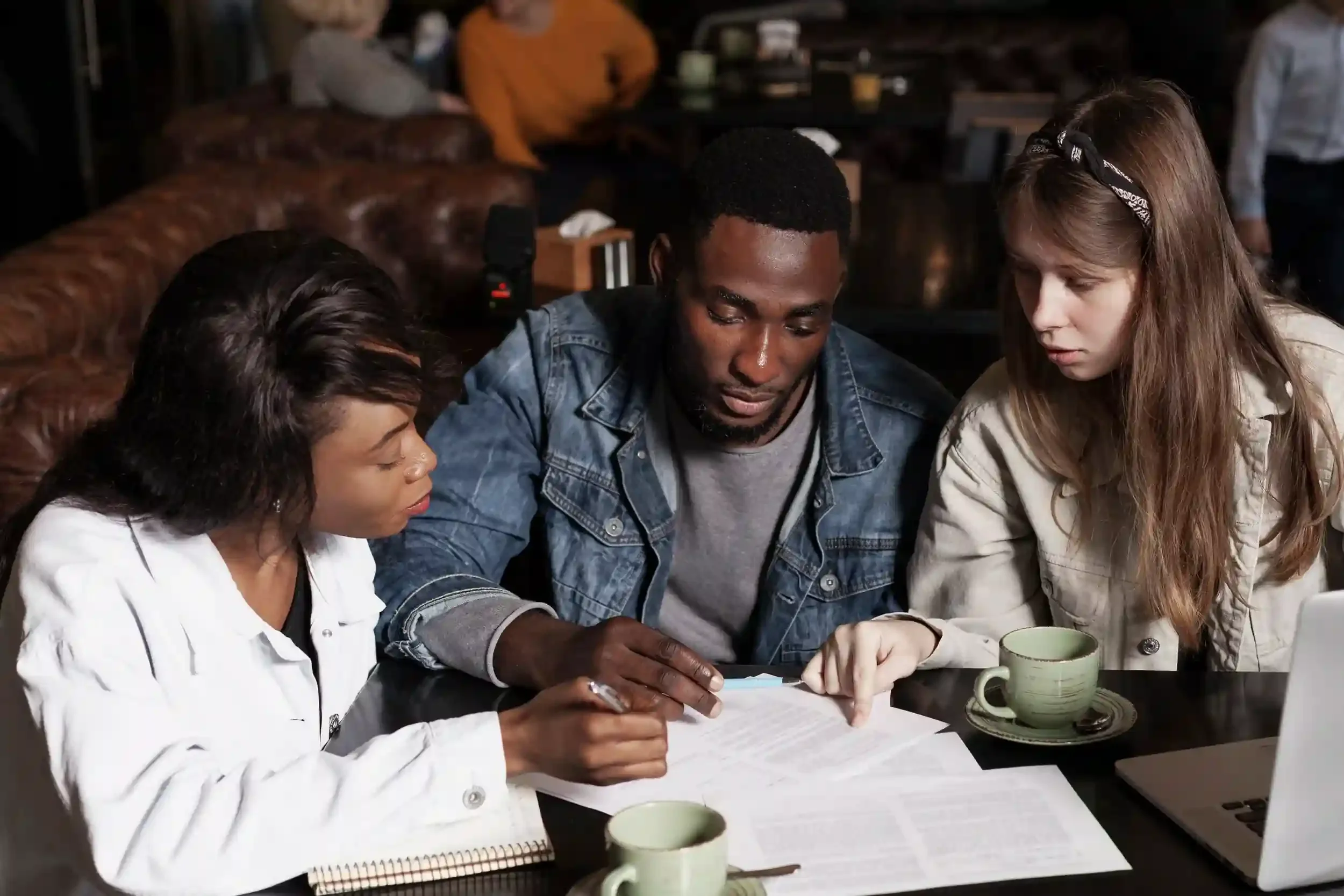 Three young people, two women and one man, sitting at a table with papers, notebooks, a laptop, and coffee cups, engaged in a discussion.