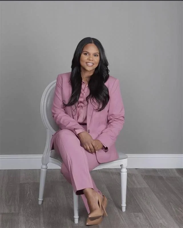 A woman with long black hair sitting on a white chair against a gray wall, wearing a pink suit and nude high heels.
