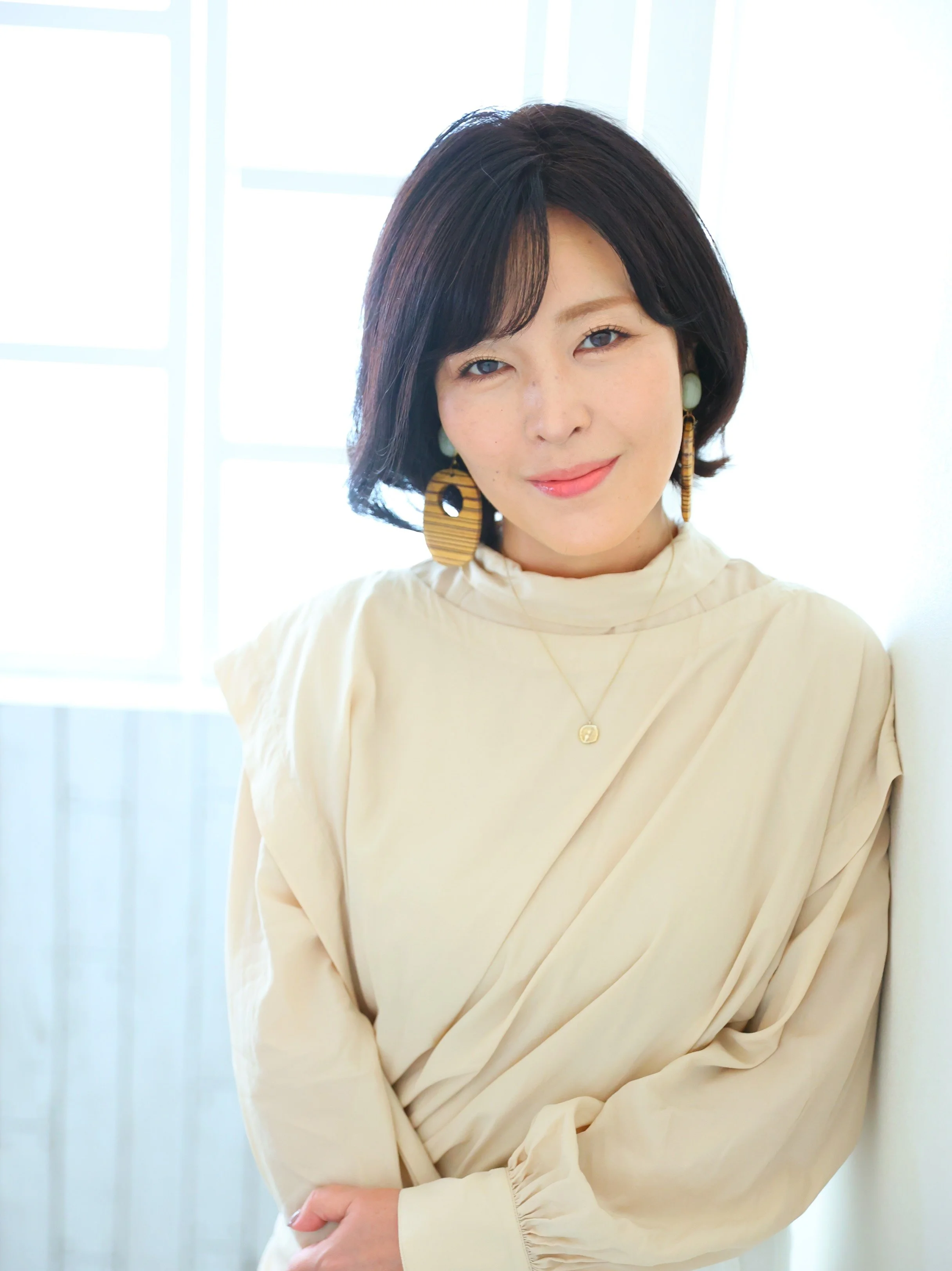 白い布のトップスを着た女性が微笑みながらカメラを見る。背景は明るい窓と白い壁。