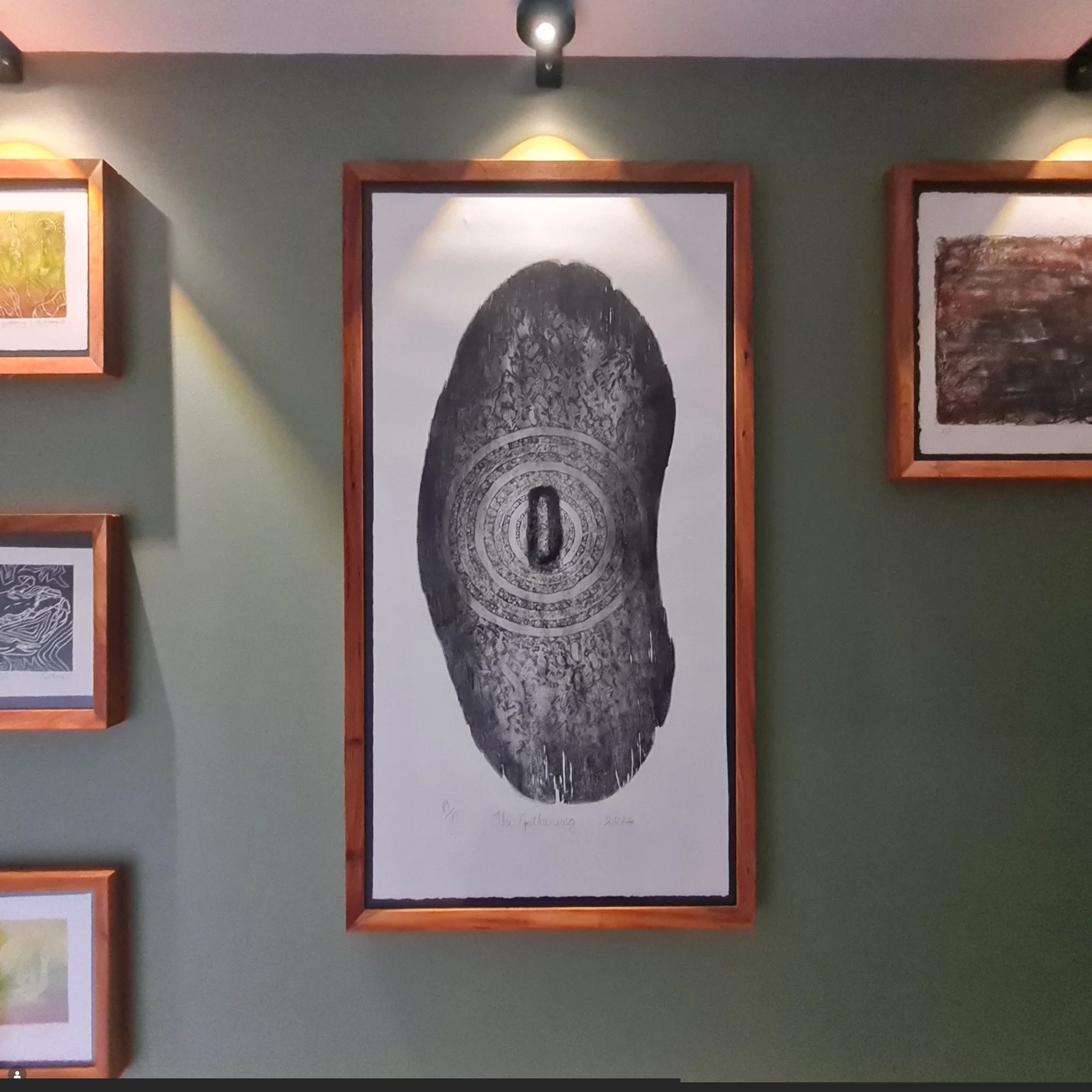 Artwork depicting a large Aboriginal shield print with intricate designs and a circular pattern in the center, framed and hung on a green wall in an art gallery.