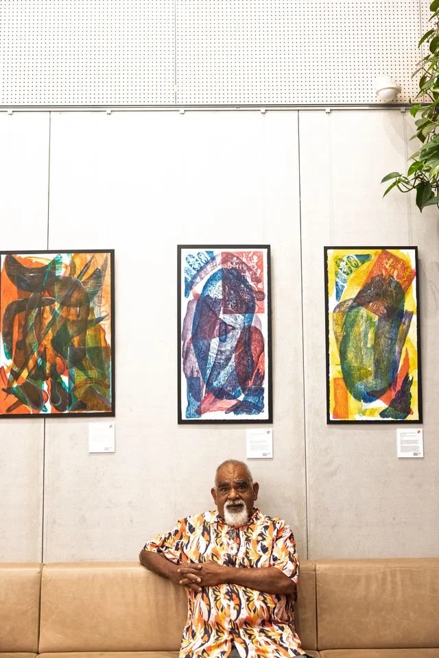 Paul sitting on a beige couch in front of abstract artwork in a gallery, wearing a colorful abstract-patterned shirt.