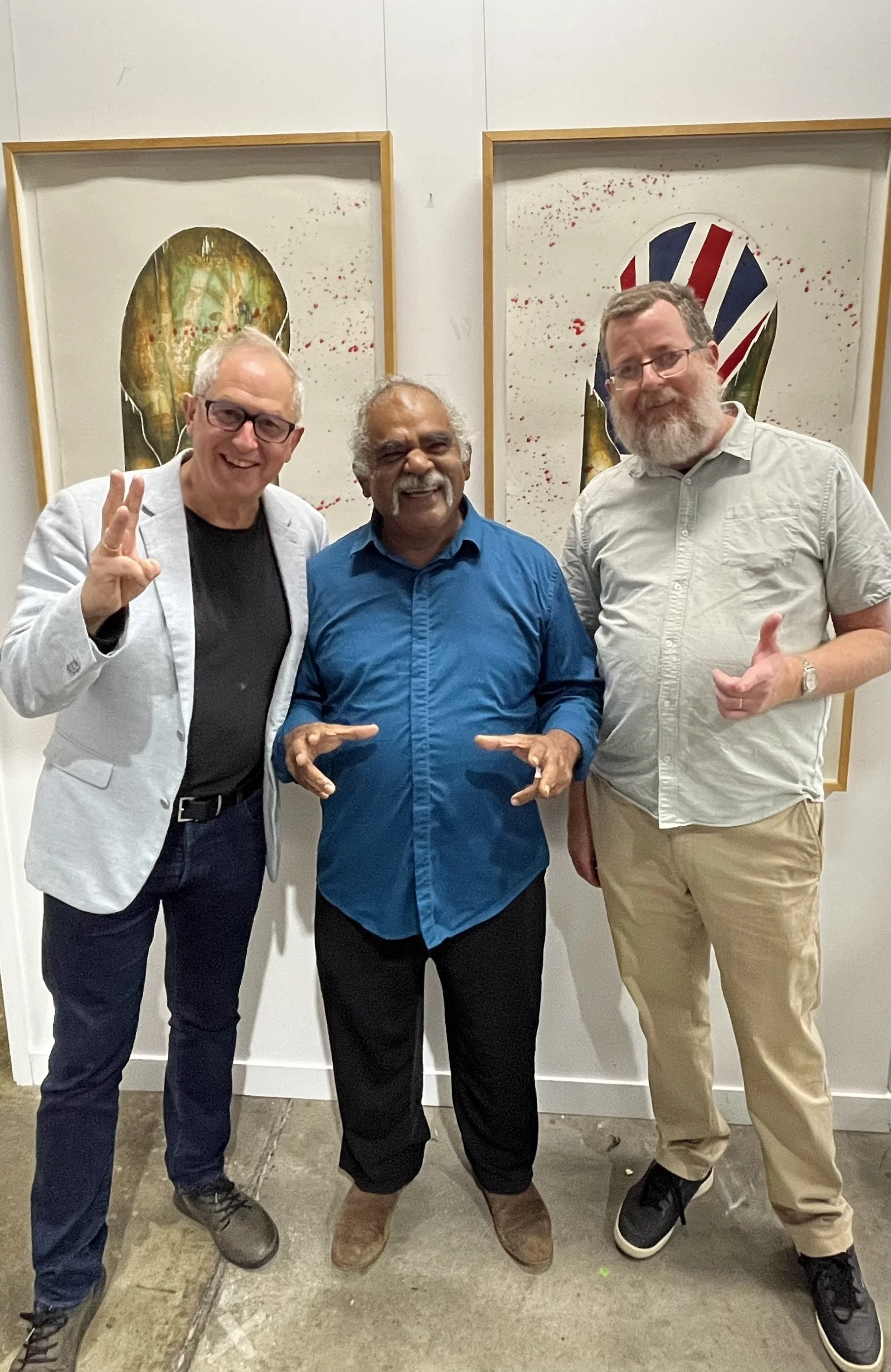 Paul with 2 other men standing in front of his artwork, smiling and making friendly hand gestures, in an art gallery.