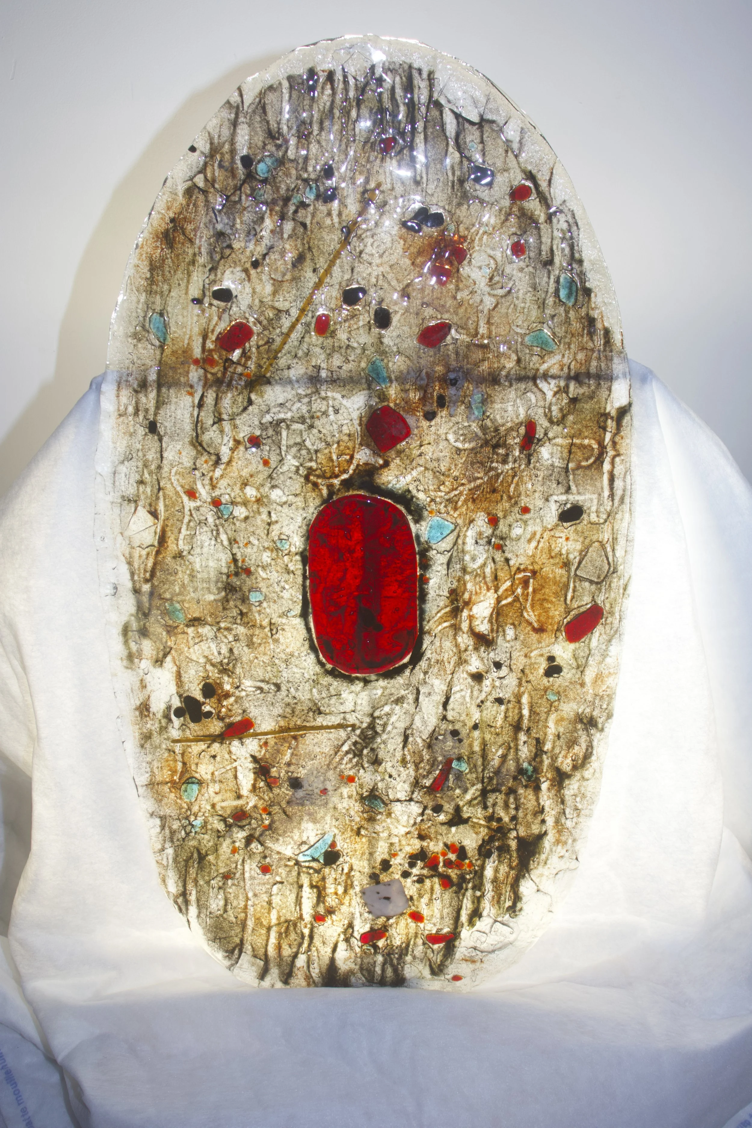 An Aboriginal shield artwork on an oval canvas featuring a large red oval in the center, surrounded by various small colored shapes and lines, with a predominantly earthy and beige background.