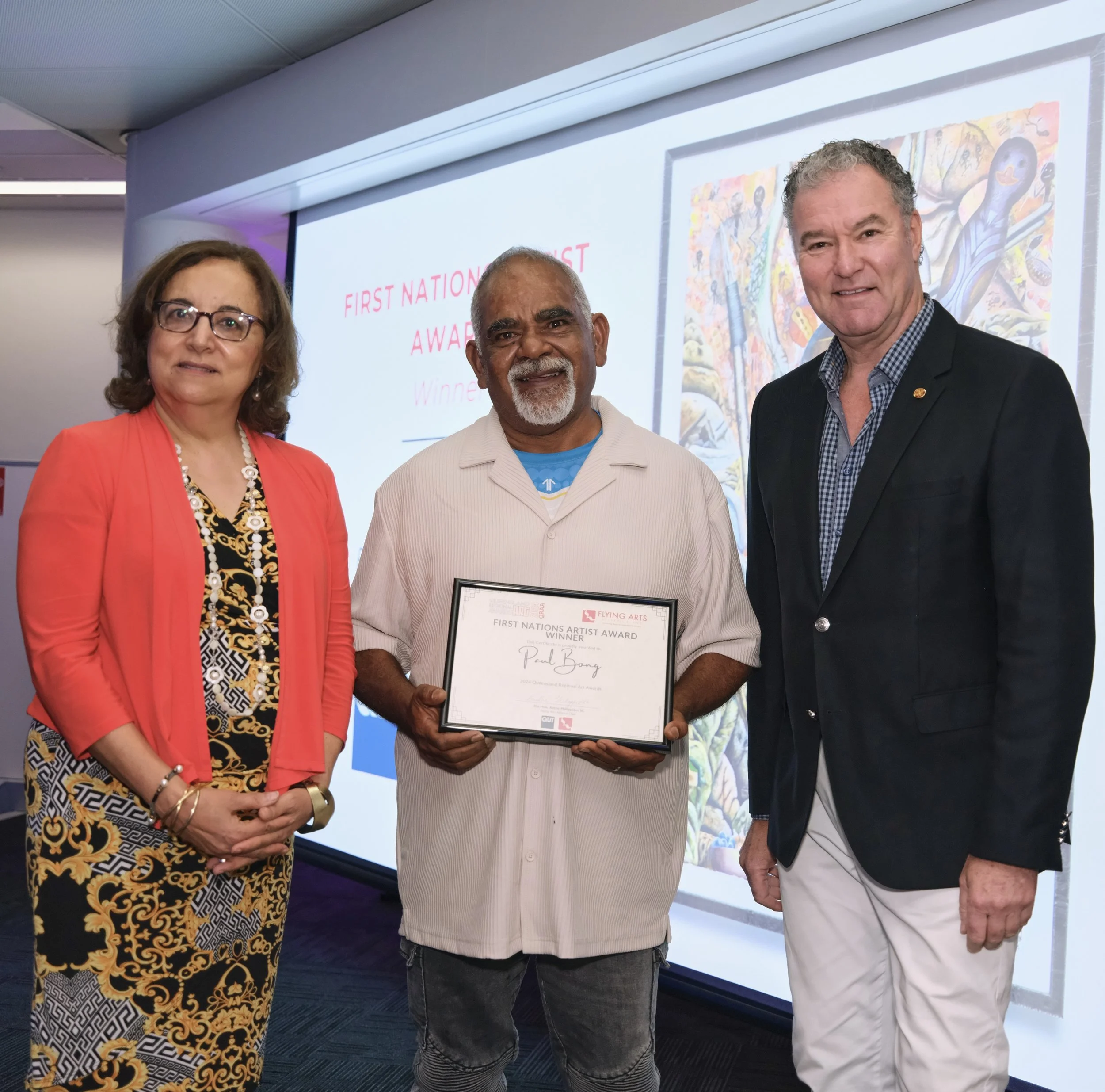 Three people standing together during an award ceremony. Paul stands in the middle, holding an award, accompanied by a woman in a red cardigan and dark dress, and a man wearing a black blazer and white trousers.