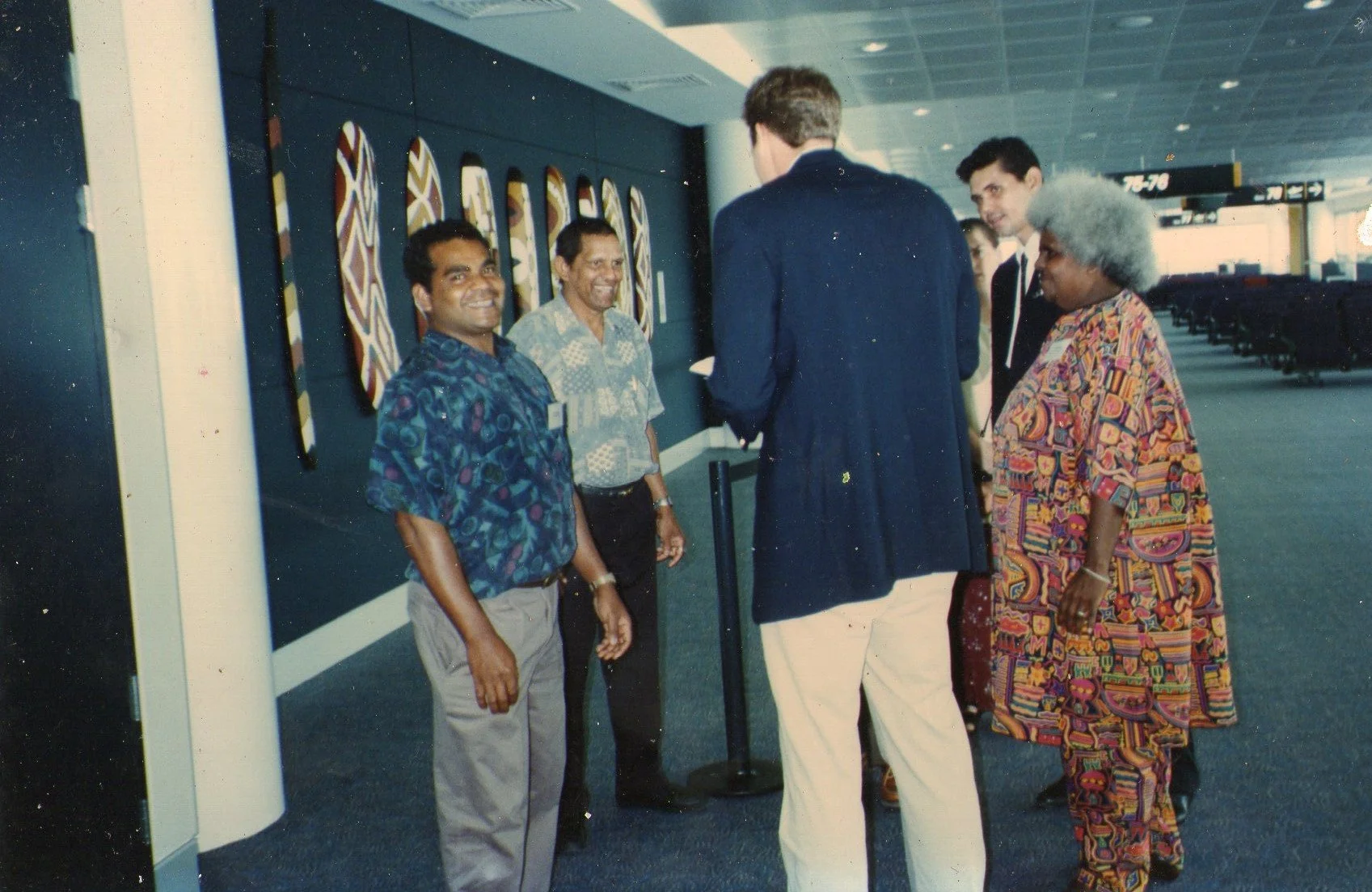 Paul with an elder and two other men standing and talking in an airport terminal, some smiling, with artwork on the wall and airport sign in the background.
