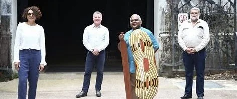 Four people standing outdoors in front of an art gallery. Paul is holding a painted aboriginal shield.