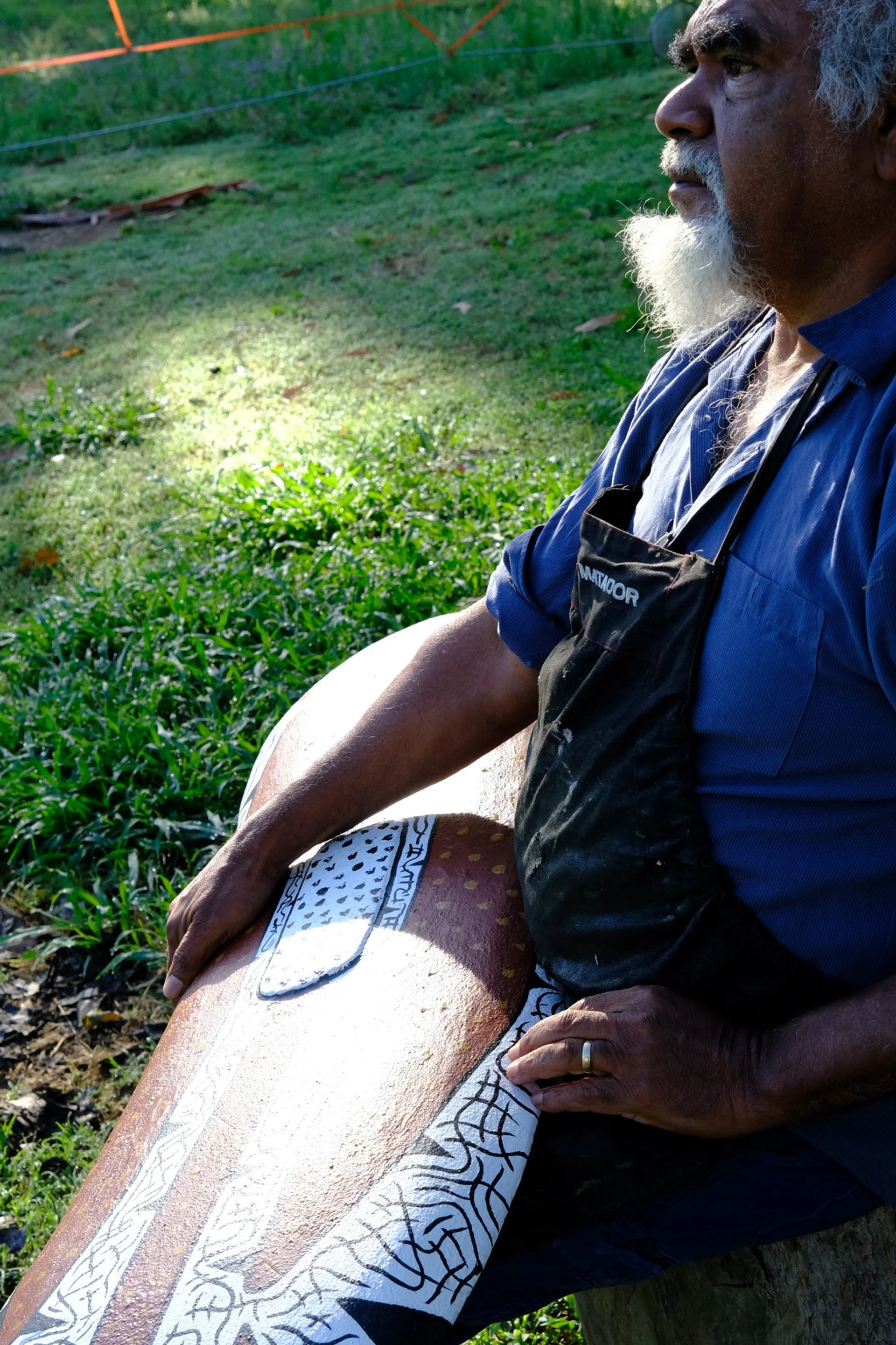 Paul Bong, wearing a blue shirt and black apron, sits outdoors on a wooden bench. He is working on a piece of wood with intricate painted patterns in black and white, with sunlight illuminating the scene and green grass in the background.