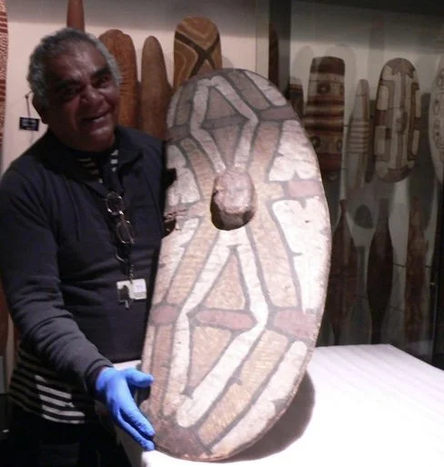 Paul holding a traditional Aboriginal shield in a museum display room with other similar artifacts in the background.