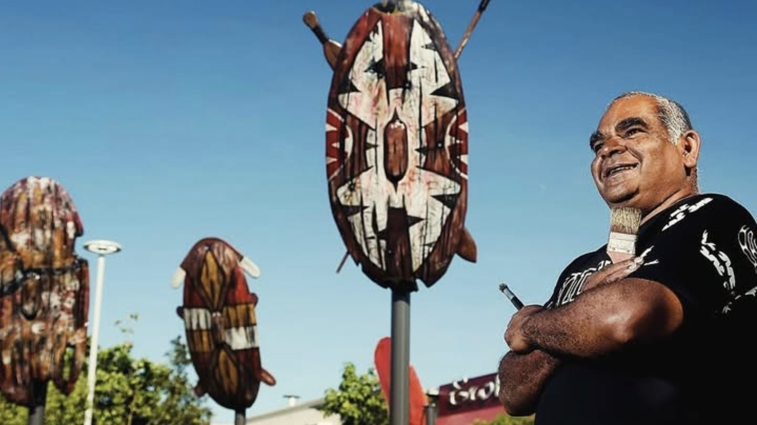 A man smiling with tribal face paint on his cheek, standing next to and behind puppets with indigenous face paintings on sticks, set against a clear blue sky.