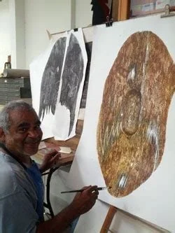 Paul painting a large, Aboriginal shield artwork on a canvas in a studio, with two other similar artworks hanging on the wall behind him.