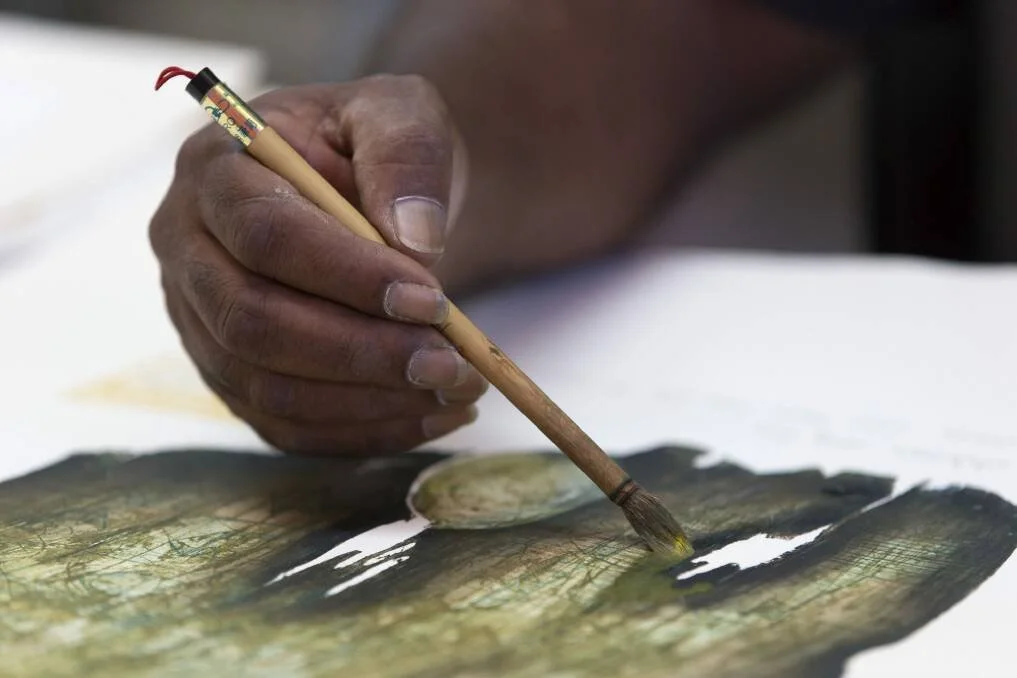 A cloes up of Paul holding a brush and painting an aboriginal shield on paper