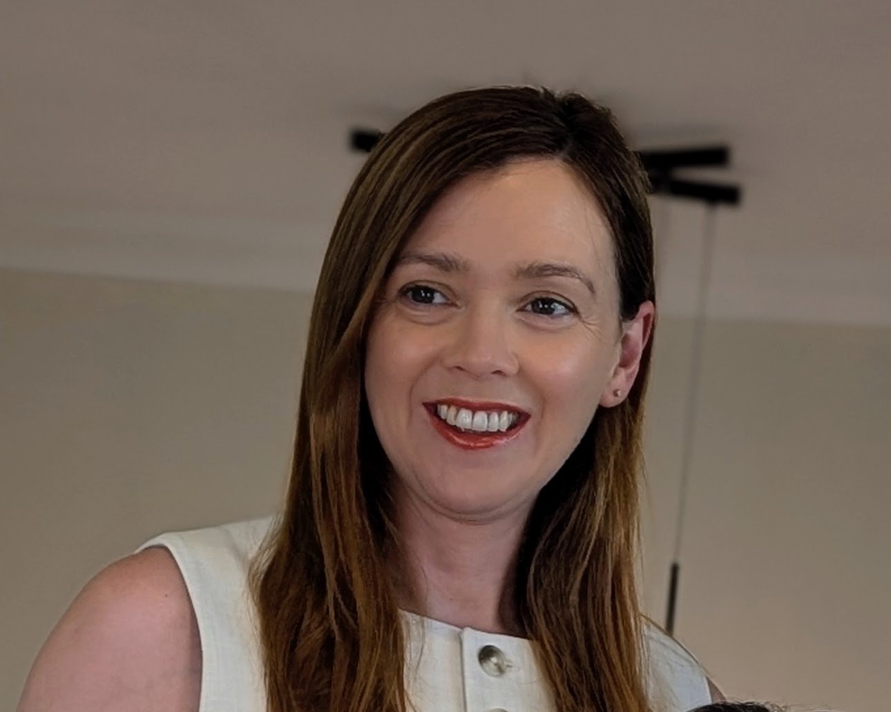 A smiling woman with long brown hair wearing a white sleeveless top indoors.