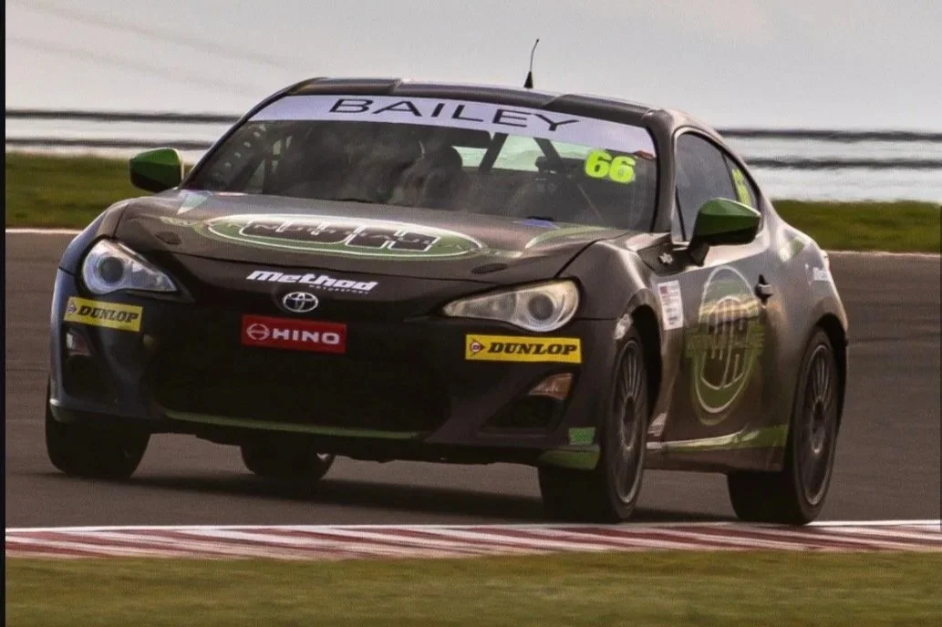 Black racing car with green accents on a racetrack, with sponsor logos including Dunlop and Hino, and the number 66 on the windshield.