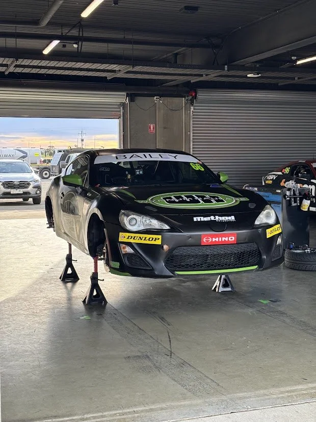 Race car on jack stands inside garage, with tires, tools, and other vehicles visible outside.