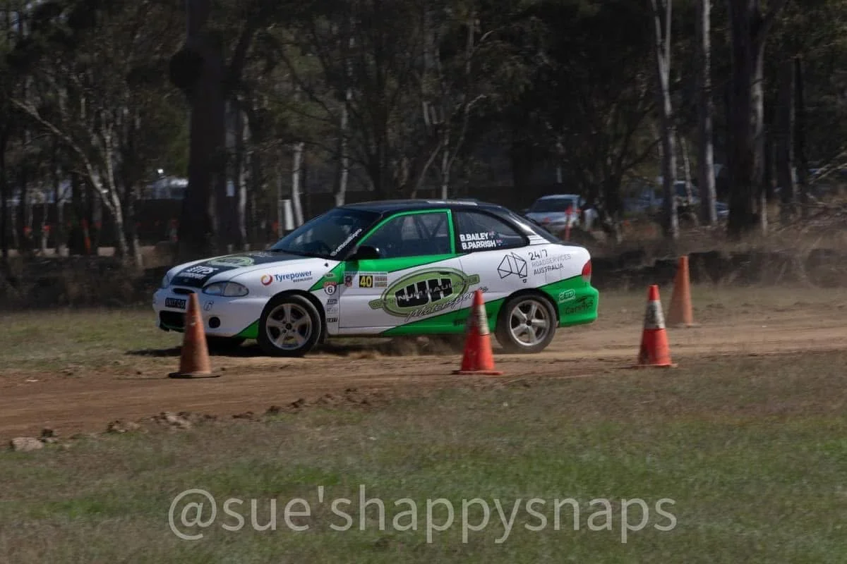 A rally car on a dirt track, surrounded by orange traffic cones, with trees in the background.