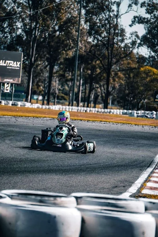 A go-kart driver wearing a helmet and racing suit driving around a track with trees in the background.
