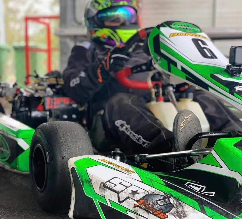 A race car driver in a colorful helmet and black racing suit sitting in a green and black open-wheel race car, holding the steering wheel.