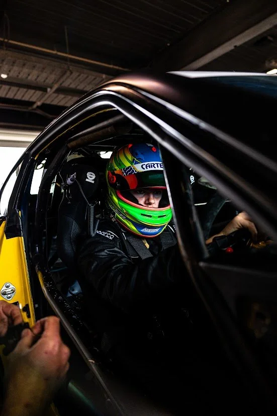 Race car driver wearing a colorful helmet sitting inside a race car in a garage.