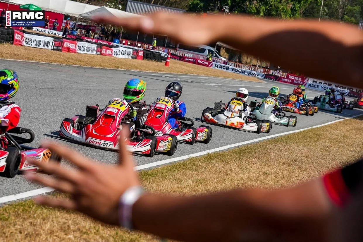 A group of go-karts racing on a track, seen through a blurred foreground hand and arm. The go-karts are colorful, with some featuring large numbers on their fronts, and are driven by racers wearing helmets and racing suits. The scene is set outdoors 