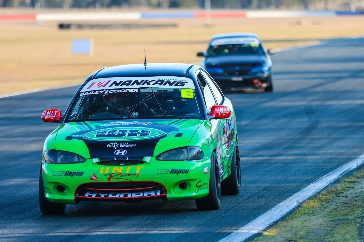 A green race car with Hyundai and UNITS Racing logos on a race track, leading a black car in the background during a race.