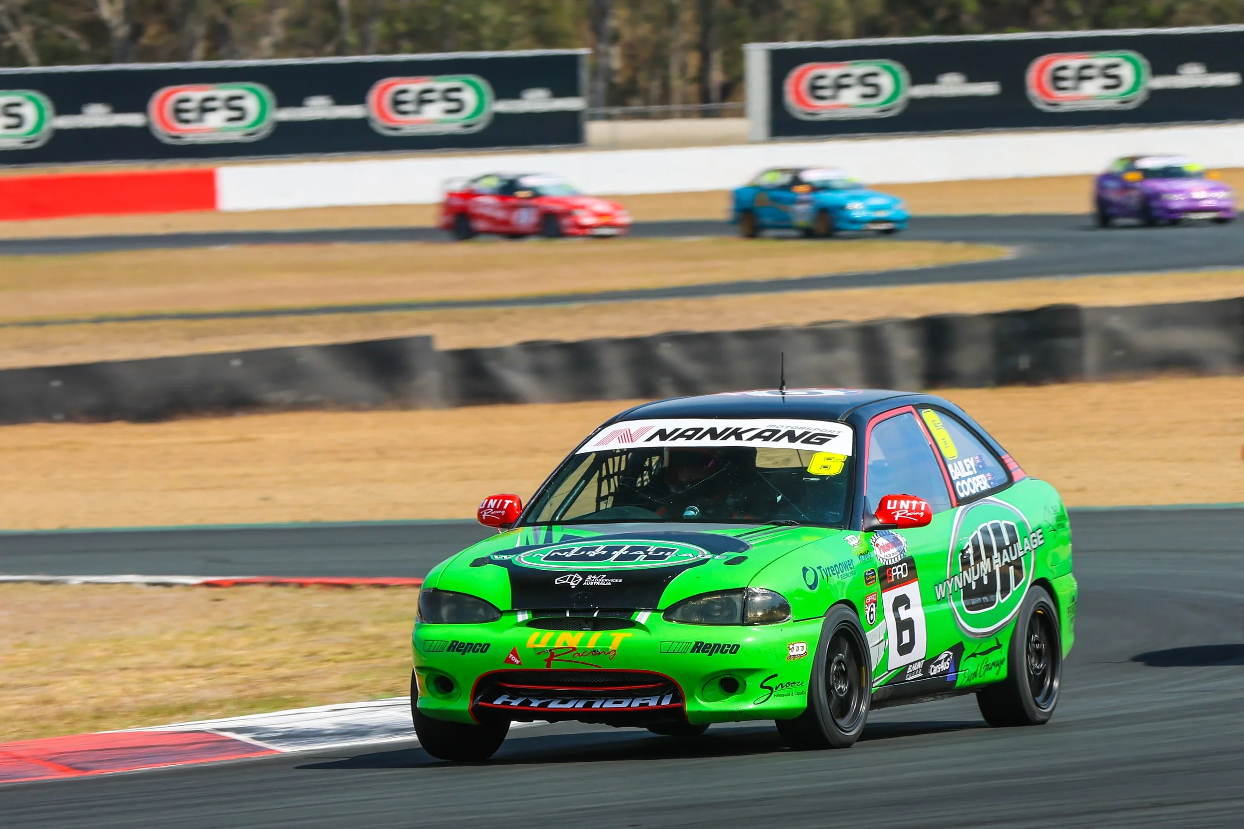 Green race car with the number 6 on its side speeding on a race track, with other race cars in the background.
