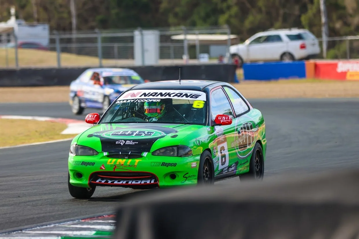 Green and black race car with the number 6 on the side, racing on a track, with a blue and white car behind it and a parking lot with vehicles in the background.