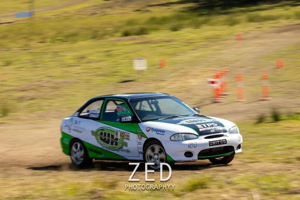 A race car moving swiftly on a dirt road during a motorsport event, with orange cones visible in the background.