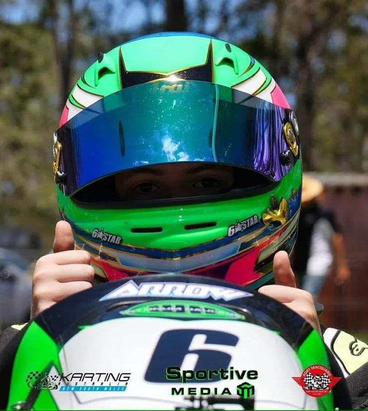 A person in a colorful racing helmet holding a karting helmet with the number 6 and various sponsor logos, standing outdoors with blurred trees in the background.