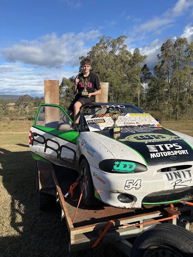 A young person sitting on top of a race car holding a trophy, with two large wooden plaques or awards behind them. The car is on a flatbed trailer, and the background features trees and a cloudy sky.