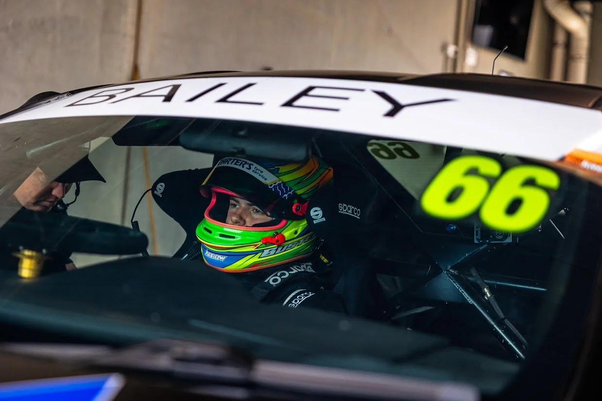 A race car driver wearing a colorful helmet sitting in a car with a person standing outside, preparing for a race. The car has the number 66 on it.