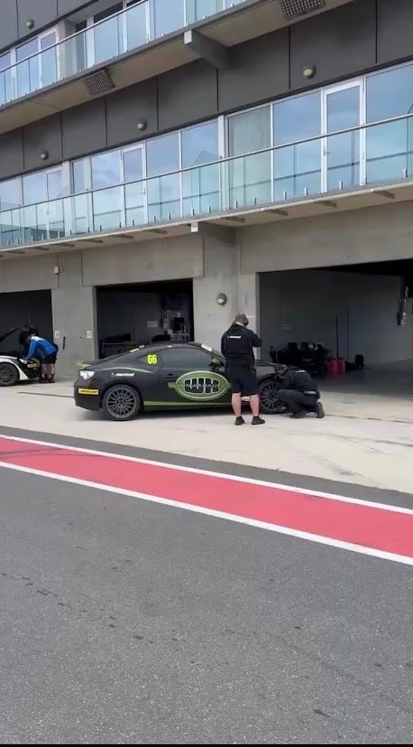 Two race car drivers preparing their black race cars in the pit garage at a racing track, with a modern apartment building in the background.
