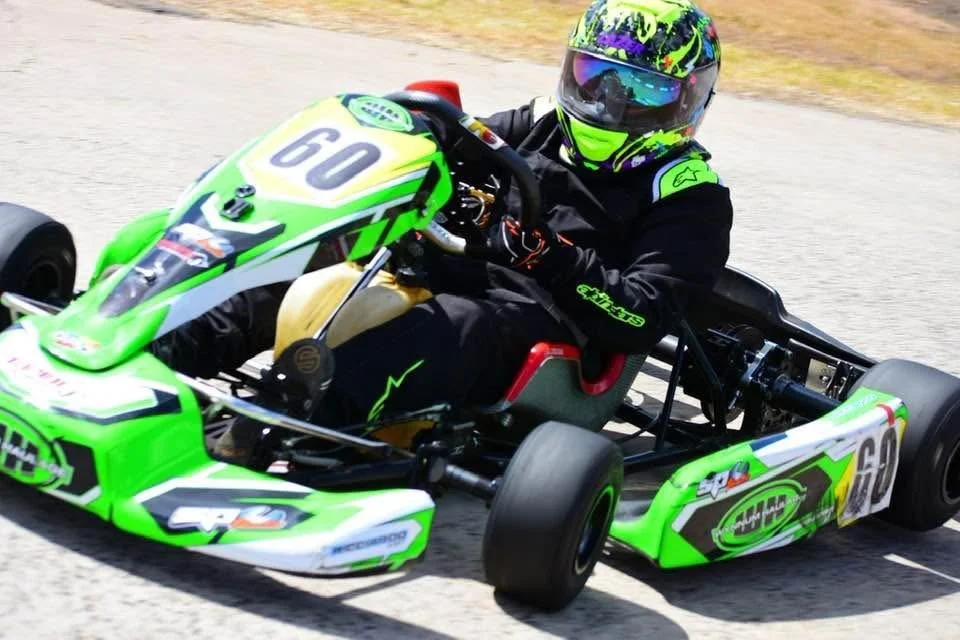 A person racing in a green go-kart on a track, wearing a black racing suit, a colorful helmet with purple, yellow, and green patterns, and reflective goggles.