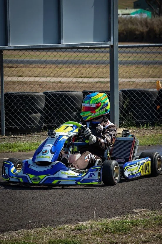 Go-kart racer wearing a colorful helmet and racing suit, sitting in a blue and yellow go-kart on a racetrack near tire barriers.