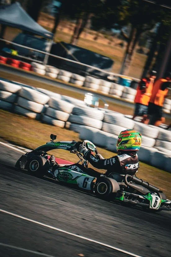 A go-kart racing on a track, with a colorful helmet and racing gear, surrounded by tire barriers and spectators.