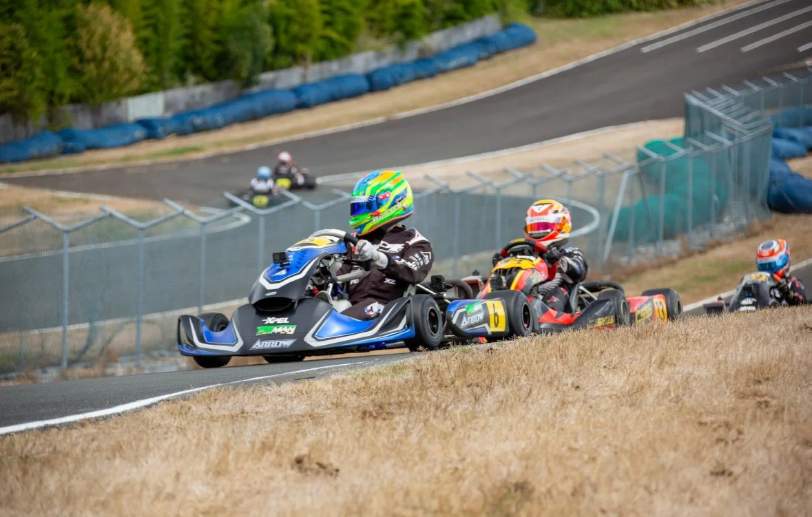 Several go-karts racing on a track with drivers wearing colorful helmets and racing suits, surrounded by a fence and barriers.