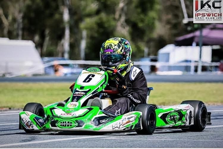 Go-kart racer dressed in black racing suit and colorful helmet driving a green and white go-kart with the number 6 on the front, on a race track with trees and tents in the background.