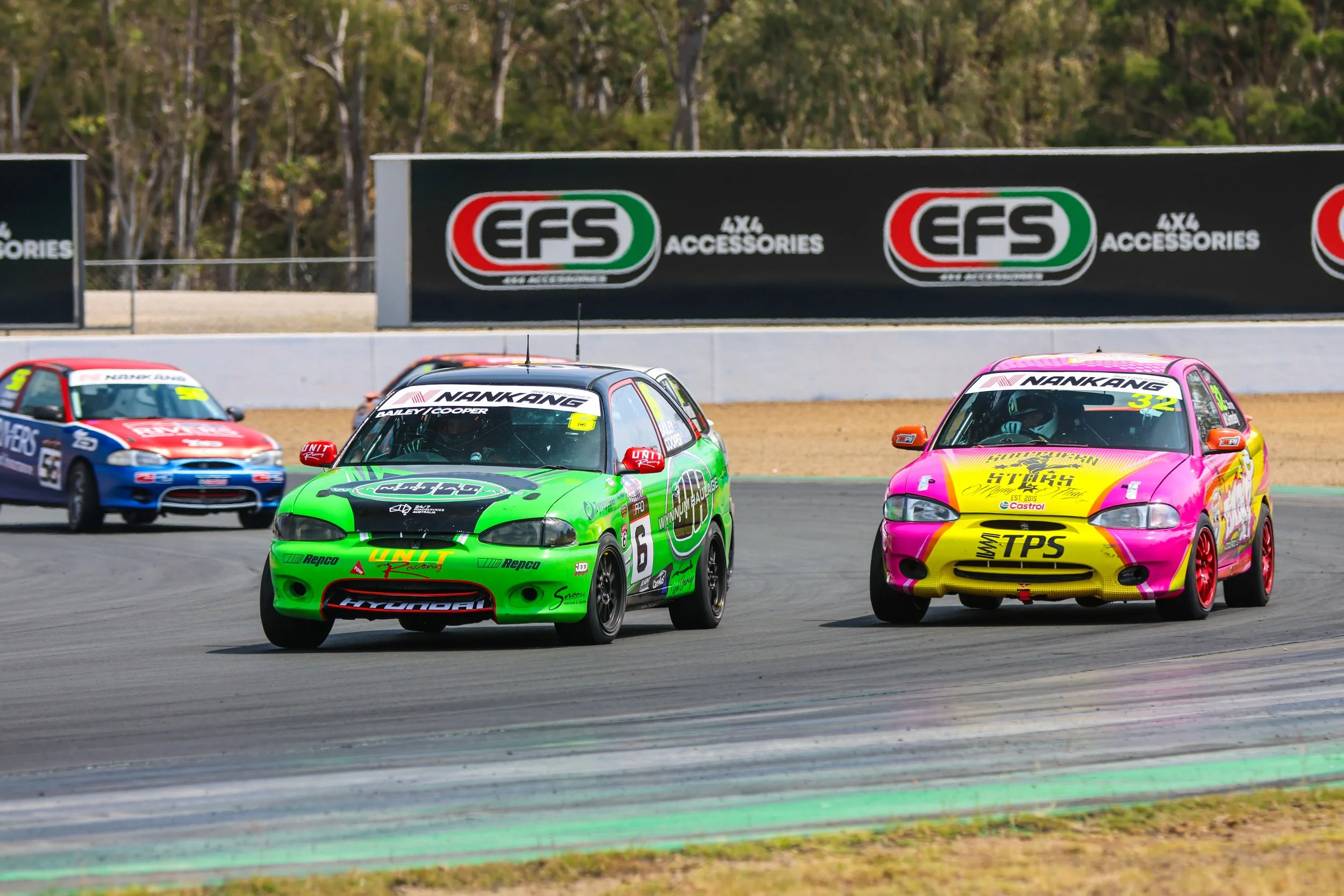 Race cars on a race track, with green, yellow, red, and blue cars, passing a black advertisement board with the logo 'EFS' in the background.